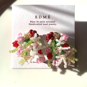 Könnte beinhalten: Ein Paar Ohrstecker bestehend aus Clustern von mehrfarbigen Glasperlen in Rot, Hellrosa, Limettengrün und klarem Weiß. Die Ohrringe sind auf einer weißen Karte befestigt, die den Text EDMÉ Handcrafted bead jewelry trägt.