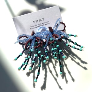 May include: A pair of beaded earrings attached to a white display card labeled EDMÉ Bijou de perle artisanal Handcrafted bead jewelry. The earrings feature loops of light blue and dark brown seed beads with dangling strands of dark blue and turquoise beads.