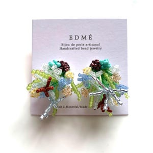 May include: A pair of handcrafted bead earrings. The earrings feature a cluster of colourful beads in shades of green, blue, yellow, brown, and silver. The earrings are on a white card with the text "EDMÉ" and "Bijou de perle artisanal Handcrafted bead jewelry".
