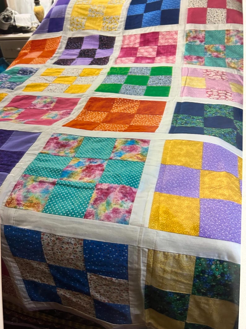 K&ouml;nnte beinhalten: Ein bunter Patchwork-Quilt mit einem Rastermuster. Der Quilt zeigt Quadrate in verschiedenen Farben und Mustern, darunter Blumenmuster, Punkte und unifarbene Stoffe. Der Quilt ist mit einem wei&szlig;en Stoff eingefasst.