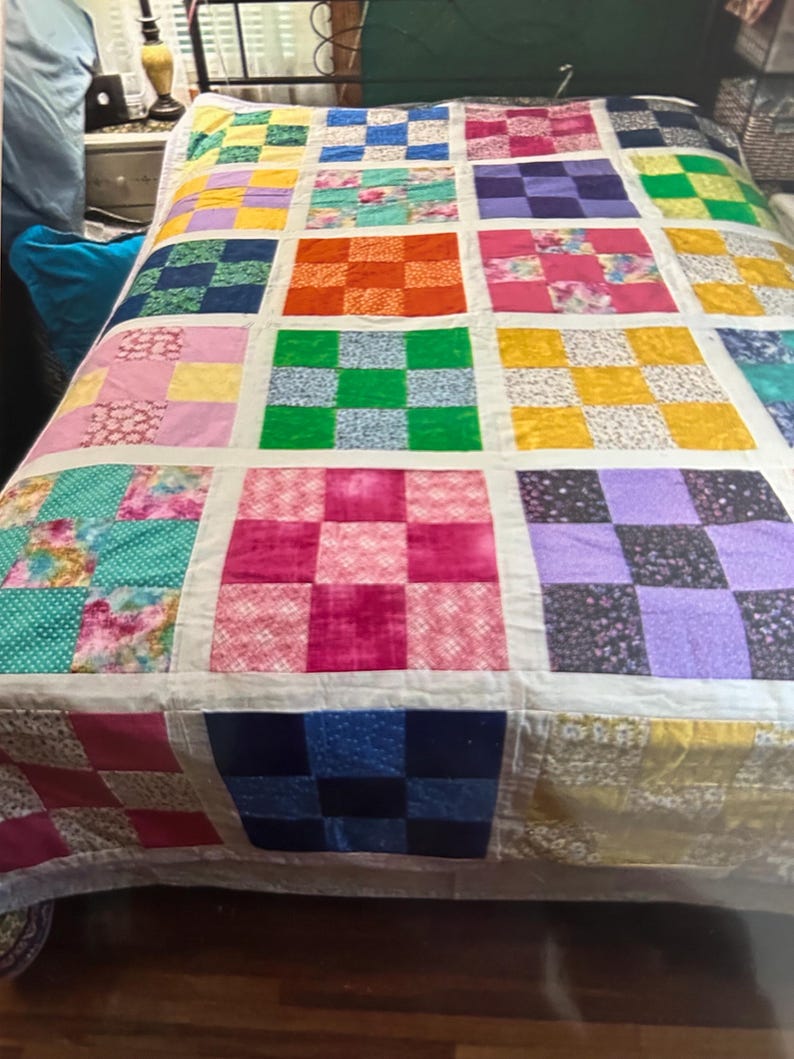 K&ouml;nnte beinhalten: Ein handgefertigter Quilt mit Patchwork-Design. Der Quilt zeigt ein Raster aus Quadraten in verschiedenen Farben, darunter Rosa, Orange, Gr&uuml;n, Blau und Gelb. Jedes Quadrat besteht aus verschiedenen gemusterten Stoffen, was eine lebendige und gem&uuml;tliche &Auml;sthetik erzeugt.