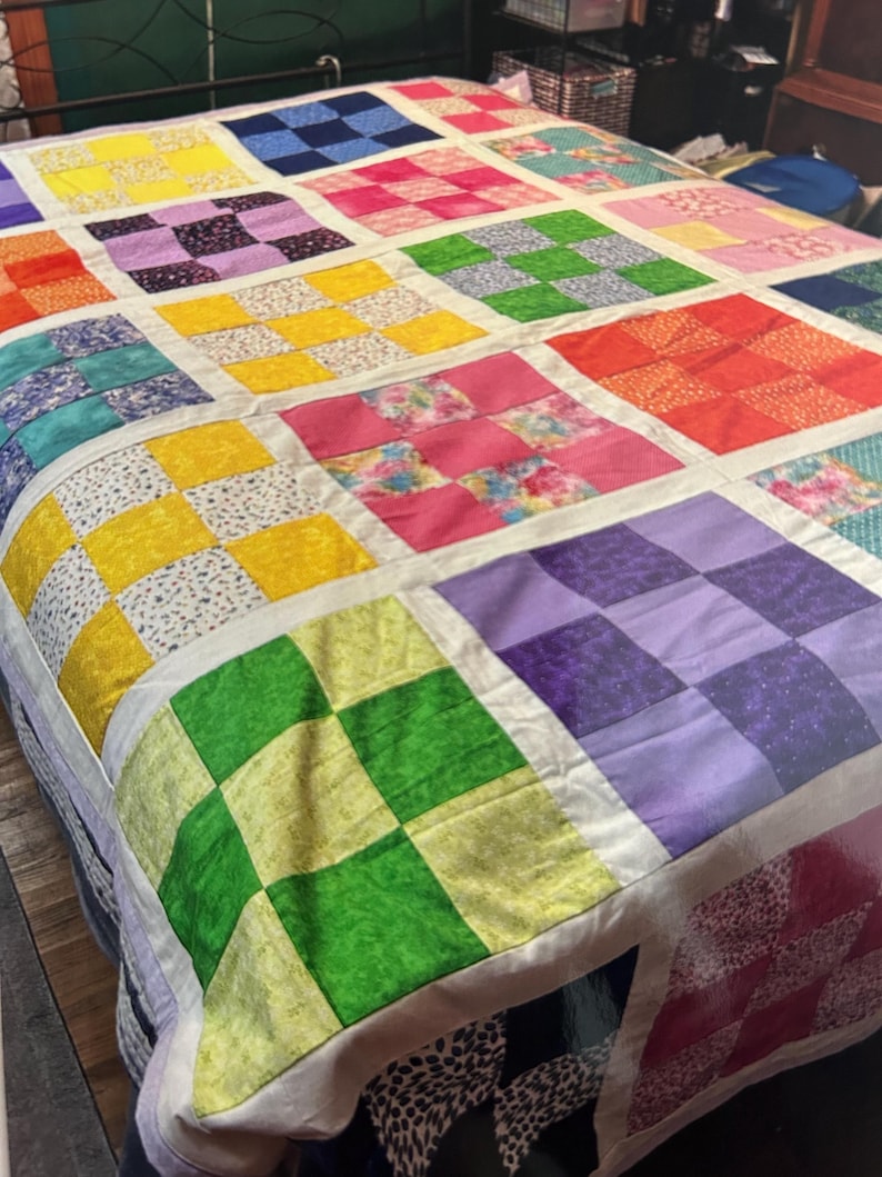 K&ouml;nnte beinhalten: Ein farbenfroher Patchwork-Quilt mit einem Rastermuster aus Quadraten in verschiedenen Farben, darunter Gelb, Rosa, Gr&uuml;n und Lila. Der Quilt liegt auf einem Bett und zeigt ein handgefertigtes, gem&uuml;tliches und lebendiges Textildesign. Die Abmessungen des Quilts sind nicht sichtbar.