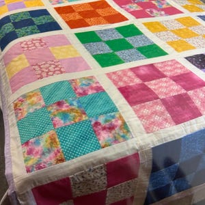 K&ouml;nnte beinhalten: Ein farbenfroher Patchwork-Quilt mit einem Rastermuster. Der Quilt zeigt Quadrate in verschiedenen Farben und Mustern, darunter Blumen-, Tupfen- und Karomuster. Der Quilt ist mit einem wei&szlig;en Stoff eingefasst und hat ein weiches, einladendes Aussehen.