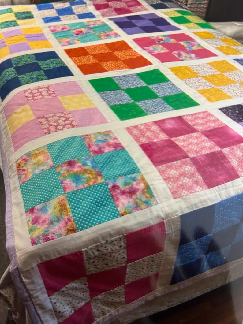 K&ouml;nnte beinhalten: Ein farbenfroher Patchwork-Quilt mit einem Rastermuster. Der Quilt zeigt Quadrate in verschiedenen Farben und Mustern, darunter Blumen-, Tupfen- und Karomuster. Der Quilt ist mit einem wei&szlig;en Stoff eingefasst und hat ein weiches, einladendes Aussehen.