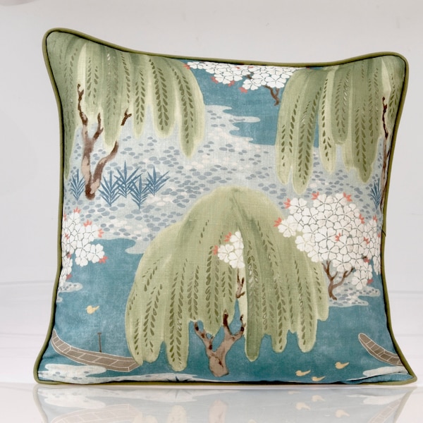 Pode incluir: Uma almofada quadrada com fundo azul e um padrão estampado de salgueiros-chorões verdes, ramos com flores brancas e pequenos barcos castanhos. A almofada é rematada com um debrum verde sólido ao longo de todo o perímetro.
