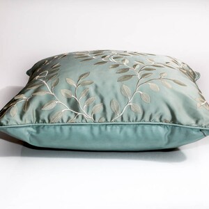 Pode incluir: Uma almofada azul claro com um design floral bordado branco. A almofada &eacute; feita de um tecido macio e luxuoso e tem um brilho sutil.