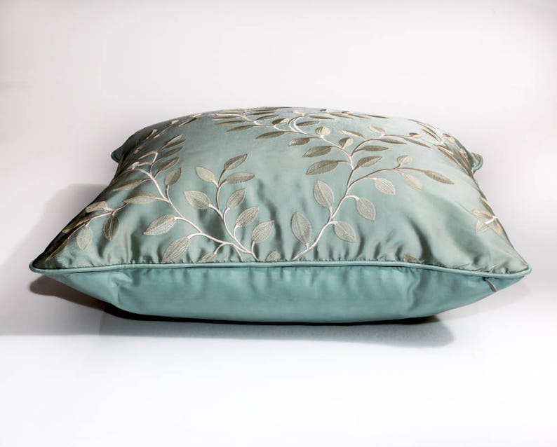 Pode incluir: Uma almofada azul claro com um design floral bordado branco. A almofada &eacute; feita de um tecido macio e luxuoso e tem um brilho sutil.