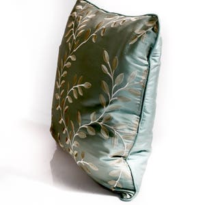 Pode incluir: Uma almofada azul claro com um padr&atilde;o floral bordado em branco e dourado.