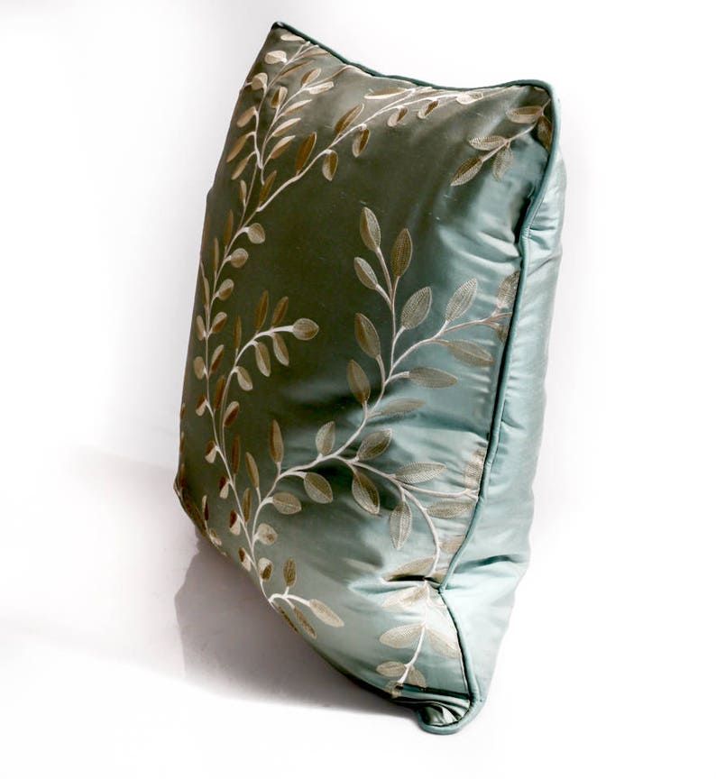 Pode incluir: Uma almofada azul claro com um padr&atilde;o floral bordado em branco e dourado.
