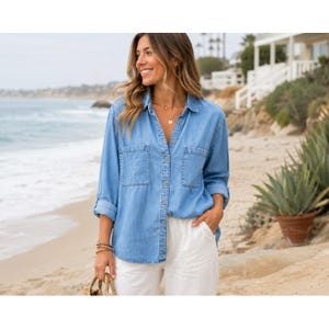May include: A light blue denim button-down shirt featuring long sleeves, a classic collar, and two chest pockets. The shirt is worn untucked over white trousers, showcasing a casual design with visible buttons running down the centre front.