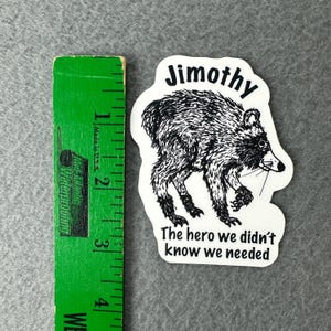 May include: A white die-cut sticker featuring a black ink illustration of a raccoon. Above the raccoon is the text Jimothy and below is the text The hero we didn't know we needed. A green ruler shows the sticker is approximately 3.5 inches tall.