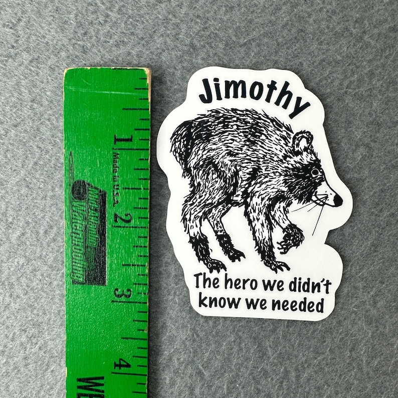 May include: A white die-cut sticker featuring a black ink illustration of a raccoon. Above the raccoon is the text Jimothy and below is the text The hero we didn't know we needed. A green ruler shows the sticker is approximately 3.5 inches tall.