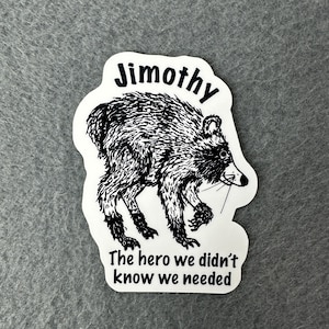 May include: A white die-cut sticker featuring a black and white ink-style illustration of a raccoon. The text "Jimothy" is printed above the animal, and the phrase "The hero we didn't know we needed" is printed below in a black sans-serif font.