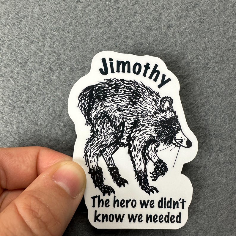 May include: A white die-cut sticker featuring a black ink illustration of a raccoon. The text "Jimothy" is printed in black above the animal, and the text "The hero we didn't know we needed" is printed in black below the raccoon illustration.