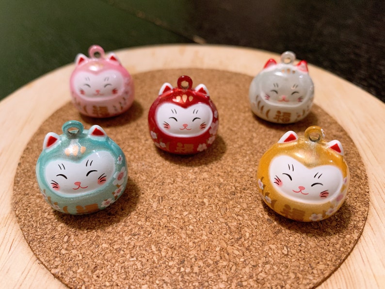 Peut inclure: Cinq figurines rondes de chat color&eacute;es avec un visage souriant et une boucle pour suspendre. Les figurines sont dans des tons de rose, rouge, vert, or et gris. Les figurines de chat sont sur une surface en bois.