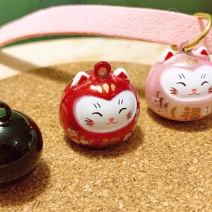Peut inclure: Trois breloques japonaises Daruma en forme de chat, rouges, roses et noires. Les breloques rouges et roses pr&eacute;sentent un motif de visage de chat souriant avec des accents dor&eacute;s. La breloque noire est une sph&egrave;re brillante.