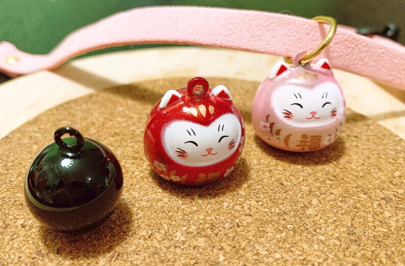 Peut inclure: Trois breloques japonaises Daruma en forme de chat, rouges, roses et noires. Les breloques rouges et roses pr&eacute;sentent un motif de visage de chat souriant avec des accents dor&eacute;s. La breloque noire est une sph&egrave;re brillante.