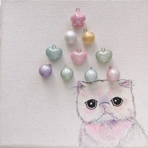 Peut inclure: Une &oelig;uvre d'art fantaisiste repr&eacute;sentant un chat blanc aux grands yeux, orn&eacute; de breloques color&eacute;es. Les breloques comprennent une &eacute;toile rose, une boule argent&eacute;e &agrave; paillettes, une boule dor&eacute;e, des breloques en forme de c&oelig;ur et rondes de diff&eacute;rentes couleurs pastel. L'&oelig;uvre est sur une toile blanche.