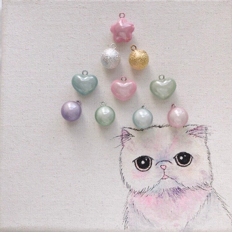 Peut inclure: Une &oelig;uvre d'art fantaisiste repr&eacute;sentant un chat blanc aux grands yeux, orn&eacute; de breloques color&eacute;es. Les breloques comprennent une &eacute;toile rose, une boule argent&eacute;e &agrave; paillettes, une boule dor&eacute;e, des breloques en forme de c&oelig;ur et rondes de diff&eacute;rentes couleurs pastel. L'&oelig;uvre est sur une toile blanche.