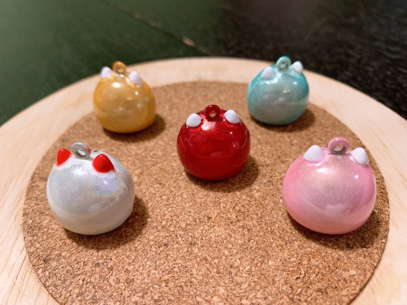 Peut inclure: Cinq breloques rondes color&eacute;es avec de petites boucles pour suspendre. Les breloques sont dans des tons or, rouge, sarcelle, rose et blanc. Chaque breloque a deux protub&eacute;rances blanches en forme d'oreille. Les breloques sont expos&eacute;es sur une surface en li&egrave;ge.