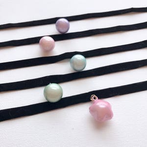 Peut inclure: Vue a&eacute;rienne de plusieurs colliers ras du cou. Chaque collier est compos&eacute; d'un ruban de velours noir avec un pendentif perle ou breloque. Les perles sont dans des tons de rose, vert et violet. Un pendentif est un charm en forme d'&eacute;toile rose.