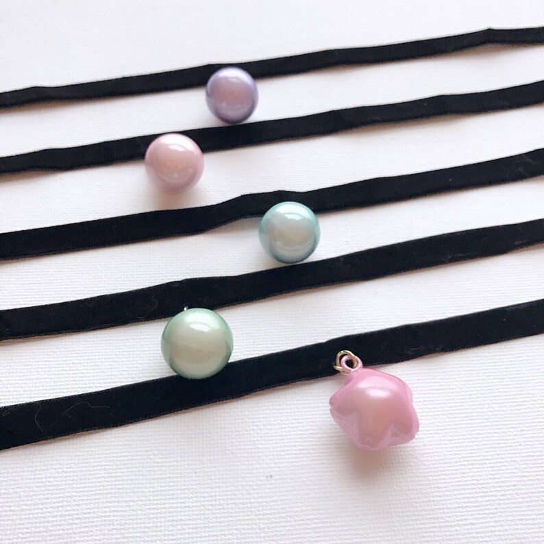 Peut inclure: Vue a&eacute;rienne de plusieurs colliers ras du cou. Chaque collier est compos&eacute; d'un ruban de velours noir avec un pendentif perle ou breloque. Les perles sont dans des tons de rose, vert et violet. Un pendentif est un charm en forme d'&eacute;toile rose.