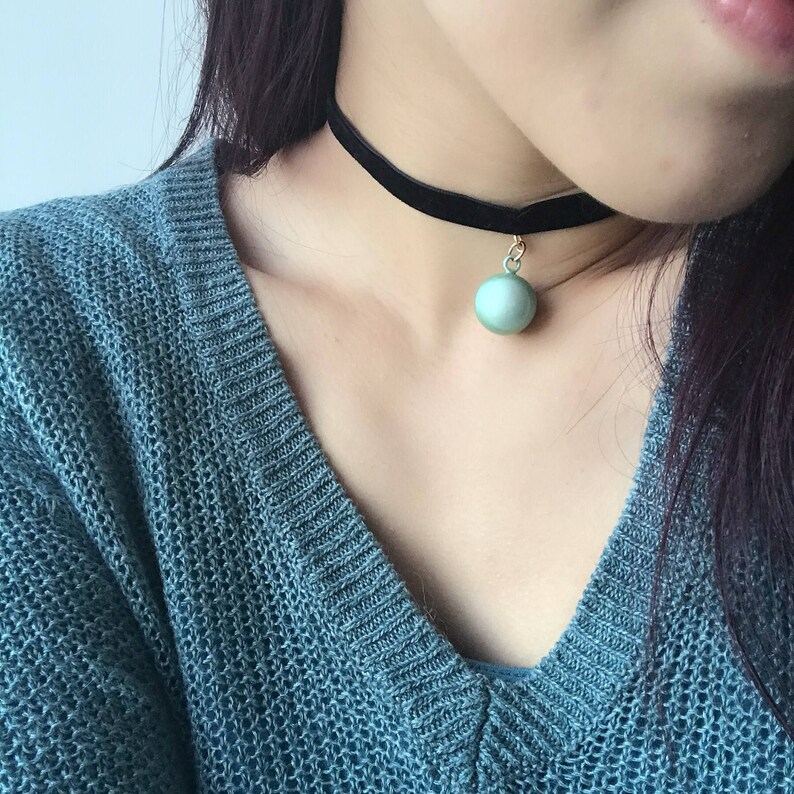 Peut inclure: Un collier ras du cou en velours noir avec un petit pendentif rond vert p&acirc;le. Le pendentif est suspendu &agrave; une cha&icirc;ne dor&eacute;e. Le collier est port&eacute; sur un pull bleu-vert.