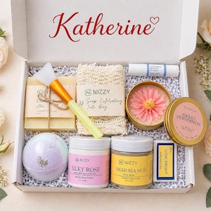 May include: A gift box filled with beauty products, including soaps, a bath bomb, body butter, face mask, and a candle. The box is labeled "Katherine" in red script. The products are arranged on white crinkle paper, with floral accents.