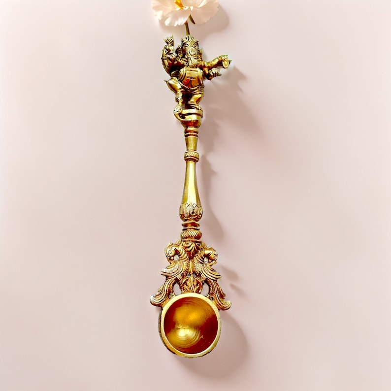 May include: A gold-colored metal spoon with an ornate design featuring a small statue of the Hindu deity Ganesha at the top. The spoon has a round bowl and a long handle.