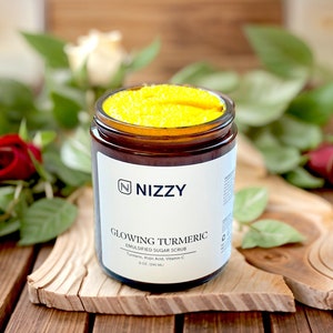 May include: A glass jar with a yellow sugar scrub labeled "Glowing Turmeric Emulsified Sugar Scrub" with the brand name "Nizzy" in a dark brown font. The jar is sitting on a wooden surface with roses in the background.