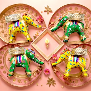 May include: Four decorative elephant figurines with colorful fabric coverings are displayed on gold and pink trays. Each elephant is carrying a small silver tray on its back. The trays are arranged in a circular pattern on a pink background.