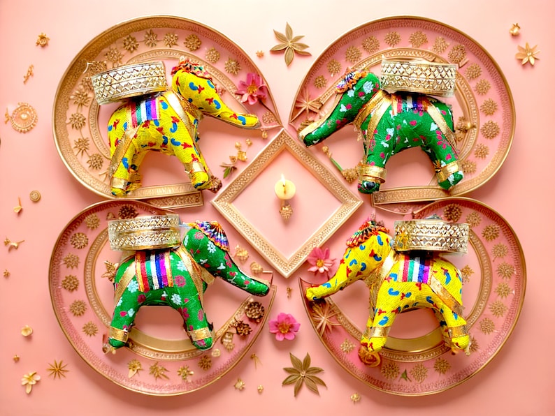 May include: Four decorative elephant figurines with colorful fabric coverings are displayed on gold and pink trays. Each elephant is carrying a small silver tray on its back. The trays are arranged in a circular pattern on a pink background.