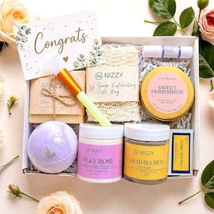 Op de afbeelding: Een geschenkdoos gevuld met zelfzorgproducten, waaronder een kaars, badbruisbal, zeep en body moisturizer. De doos bevat een kaart met "Congrats". De producten zijn gerangschikt met rozen.