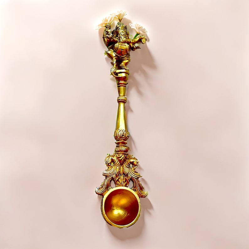 May include: A gold-colored metal diya with a decorative design featuring a deity figure and floral accents. The diya has a round bowl for holding oil or ghee.