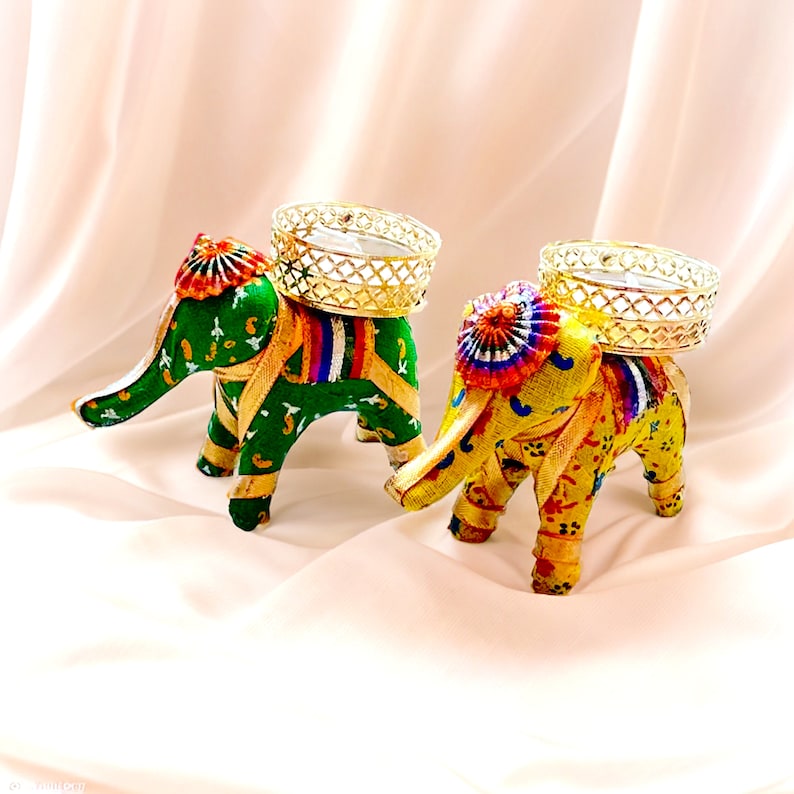 K&ouml;nnte beinhalten: Zwei dekorative Elefanten-Kerzenhalter mit aufwendigen Designs. Die Elefanten sind in leuchtenden Farben bemalt, darunter Gr&uuml;n, Gelb, Rot und Blau. Jeder Elefant hat einen goldfarbenen Metallk&auml;fig auf dem R&uuml;cken, perfekt f&uuml;r die Aufnahme einer Teelichtkerze.