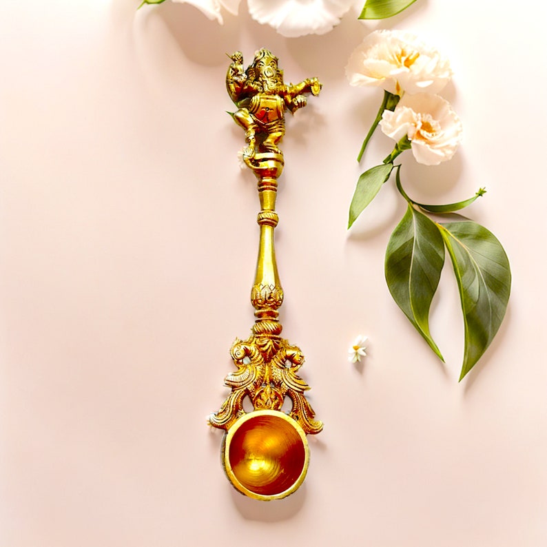May include: A gold-colored metal spoon with a detailed design featuring a figurine of the Hindu deity Ganesha. The spoon has a round bowl and a long handle with intricate carvings. The spoon is set against a light pink background with white flowers and green leaves.