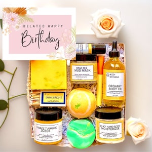 May include: A gift box filled with bath and body products, including a yellow bar of soap, a jar of orange turmeric scrub, a jar of dead sea mud mask, a bottle of organic body oil, a jar of silky hand and body moisturizer, a bath bomb, and a pack of matches. The box is decorated with a pink card that says "Belated Happy Birthday".