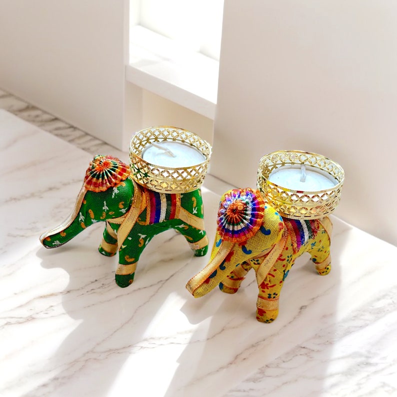 K&ouml;nnte beinhalten: Zwei dekorative Elefanten-Kerzenhalter mit goldenen Kerzenbechern aus Metall. Die Elefanten sind aus Stoff und haben bunte Muster. Ein Elefant ist gr&uuml;n und der andere ist gelb.