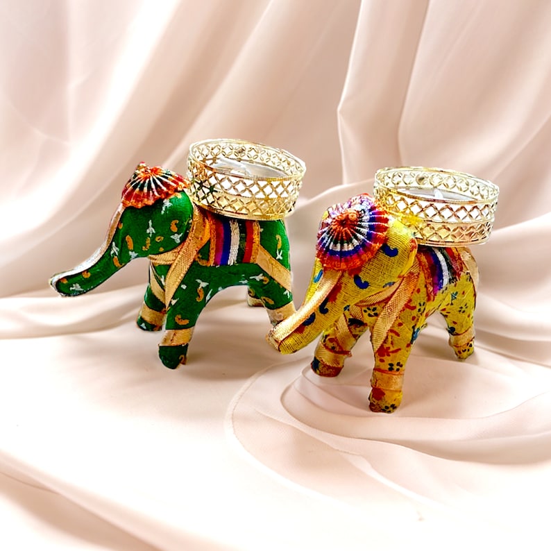 K&ouml;nnte beinhalten: Zwei dekorative Elefanten-Kerzenhalter mit aufwendigen Designs. Die Elefanten sind aus Stoff und haben einen goldenen Besatz. Jeder Elefant hat einen Kerzenhalter aus Metall auf dem R&uuml;cken.