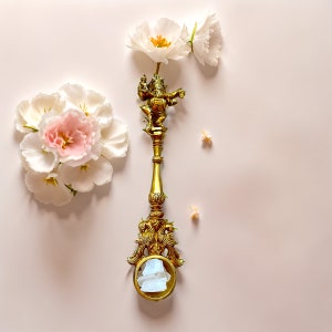May include: A gold-toned metal decorative stand with a round top and three legs. The stand features a small, ornate figure of a deity at the top. White flowers are arranged around the stand.