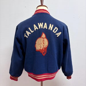 Peut inclure: Veste de style universitaire bleu marine avec le mot "TALAWANDA" en lettres blanches sur le dos. Un écusson rouge et or est centré en dessous. La veste a des poignets et un col rayés rouges et or.