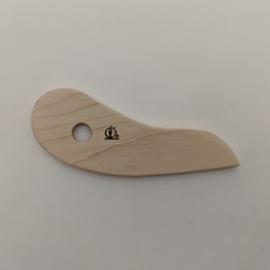May include: A light-colored wooden pottery rib tool with a curved shape and a hole. The tool has a small black logo on the surface. The tool is designed for shaping and smoothing clay in pottery.