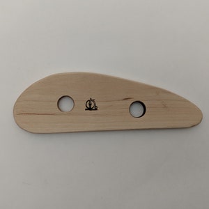 May include: A light-colored wooden pottery rib tool with two circular holes and a small black logo. The tool has a curved, ergonomic shape, ideal for shaping and smoothing clay. The wood grain is visible.