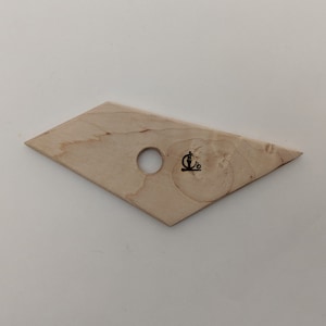 Op de afbeelding: Een ruitvormige houten pottenbakkersrib met een centraal gat en een klein zwart logo. De tool is gemaakt van lichtgekleurd hout en wordt gebruikt voor het vormen van klei. De rib is ongeveer 13 cm lang.