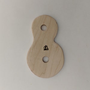 May include: A light-colored wooden tool with an hourglass shape, featuring two circular holes and a small black logo. The tool is designed with rounded edges and a smooth surface, ideal for crafting or woodworking projects.