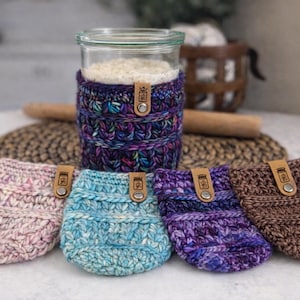 May include: A glass jar with a colorful knitted cozy, surrounded by five matching cozies in shades of pink, blue, purple, and brown. Each cozy has a leather tag with a button. The items are on a white surface.