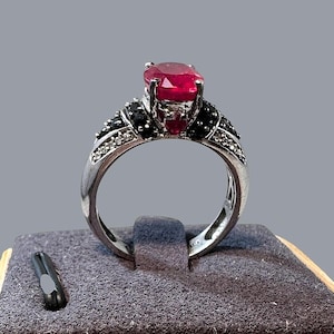 May include: A silver-toned ring featuring a large, oval-cut, deep red center stone held in a prong setting. The band is accented with smaller, dark black stones and tiny, clear white stones arranged along the shoulders of the metal setting.