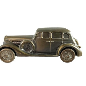 May include: A vintage-style metal car bank, painted in a dark bronze color. The car has a detailed design with a rounded body, four wheels, and a windshield.