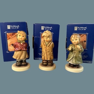 May include: Three Hummel figurines of children in winter attire, each standing on a round base. The figurines are in front of blue boxes with the M.J. Hummel Club logo. The children wear coats and hats in various colors, including red, tan, and green.