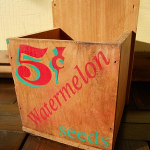 May include: A wooden box with the words "5&cent; Watermelon Seeds" painted on it in red and green.